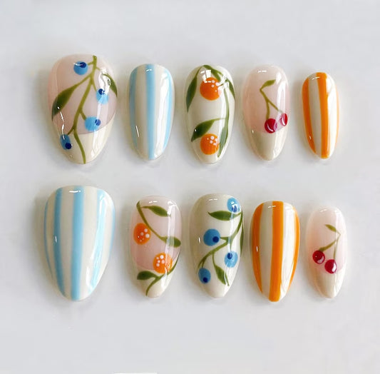Hand-painted luxury press-on nails featuring dainty botanical illustrations of oranges, cherries, and blueberries on a cream base, interspersed with sky blue and orange vertical stripes